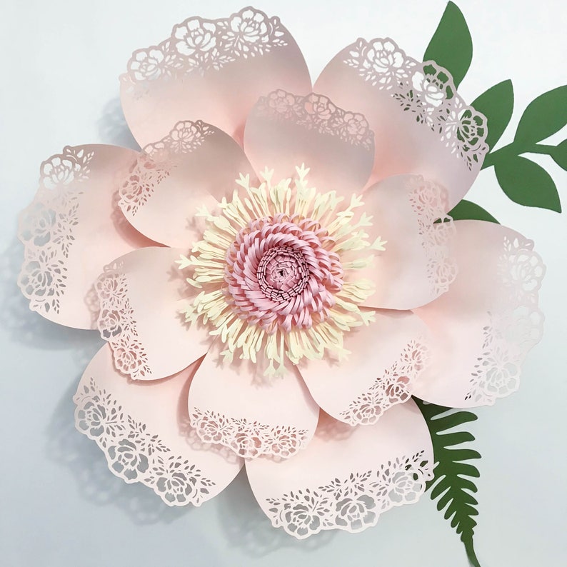 K&ouml;nnte beinhalten: Eine gro&szlig;e, rosafarbene Papierblume mit filigranen Spitzendetails und einem gelben und rosafarbenen Zentrum. Die Blume ist von gr&uuml;nen Bl&auml;ttern umgeben.