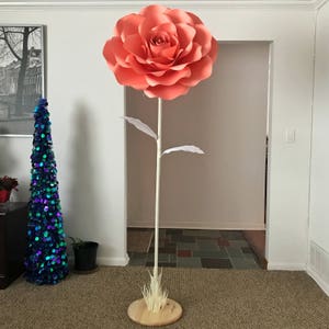 May include: A large, coral-colored paper rose on a white stem with white paper leaves. The rose is standing on a wooden base with a layer of dried grass.
