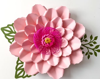 May include: A large pink paper flower with a smaller pink and yellow flower in the center. The flower is surrounded by green paper leaves.