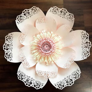 K&ouml;nnte beinhalten: Eine gro&szlig;e, rosafarbene Papierblume mit einem zarten Spitzenmuster an den R&auml;ndern. Die Mitte der Blume ist ein mehrschichtiges Papierdesign in Gelb und Rosa.