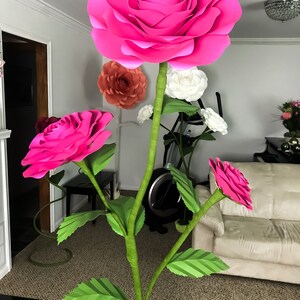 May include: A large pink paper flower with smaller pink paper flowers and green leaves on a stem. The flowers are arranged in a bouquet.