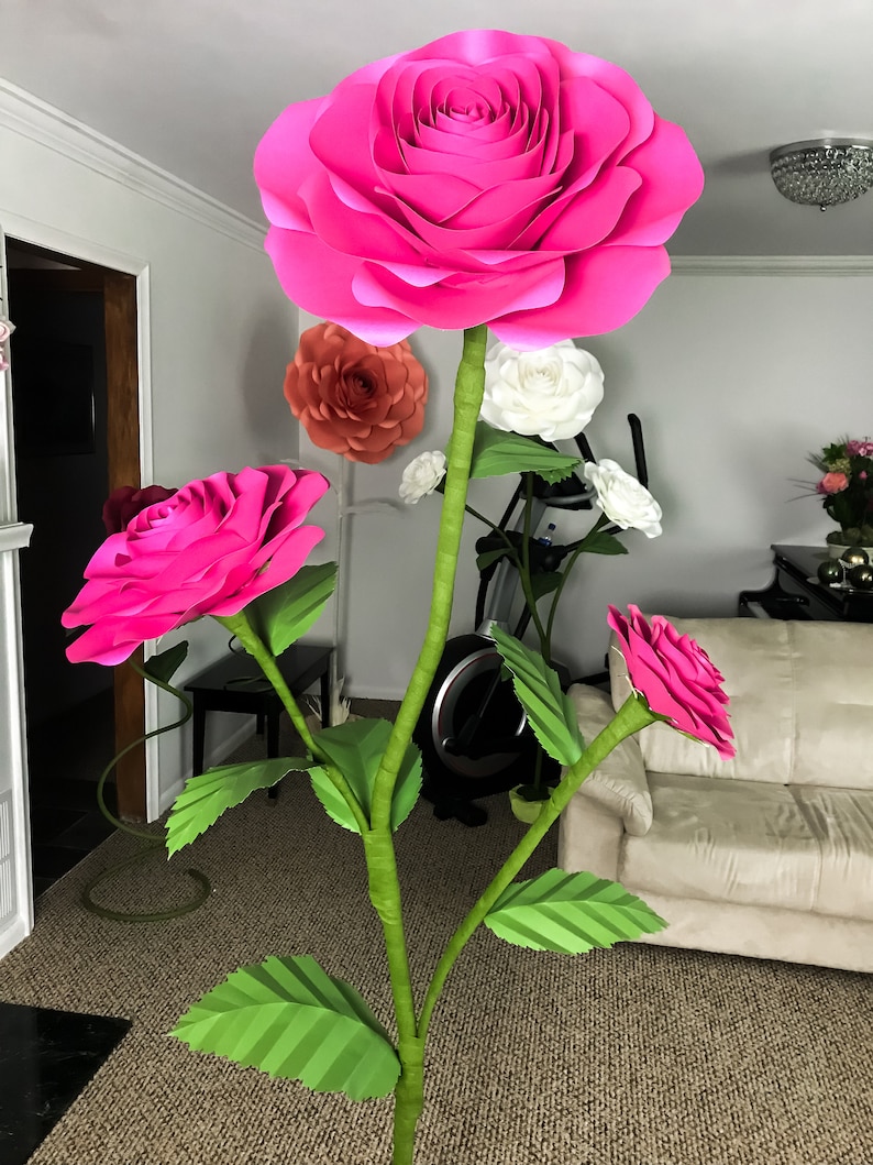 May include: A large pink paper flower with smaller pink paper flowers and green leaves on a stem. The flowers are arranged in a bouquet.