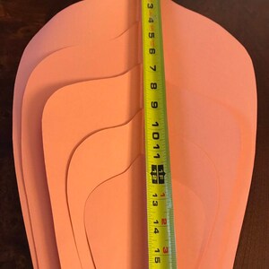 May include: Five peach-colored paper flower petals, each with a rounded top and a curved bottom, are stacked on top of each other. A yellow measuring tape is placed next to the petals, showing a measurement of 16 inches.