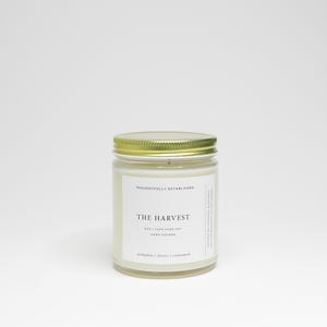 May include: A glass jar candle with a gold lid. The label reads "The Harvest" with scent notes of pumpkin, cloves, and cinnamon. The candle is 4 oz and made of 100% pure soy wax.