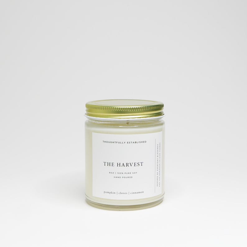 May include: A glass jar candle with a gold lid. The label reads "The Harvest" with scent notes of pumpkin, cloves, and cinnamon. The candle is 4 oz and made of 100% pure soy wax.