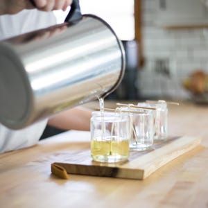 May include: A person pouring liquid from a stainless steel pitcher into clear glass jars with wicks. The jars are on a wooden board. The liquid is yellow. The background is a kitchen.