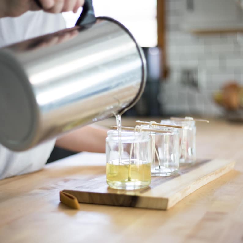 May include: A person pouring liquid from a stainless steel pitcher into clear glass jars with wicks. The jars are on a wooden board. The liquid is yellow. The background is a kitchen.