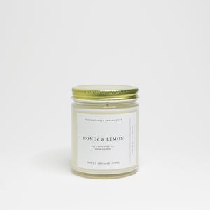 May include: A glass jar candle with a gold lid. The label reads "HONEY & LEMON" and "8oz | 100% PURE SOY | HAND POURED". The candle is white with a gold lid. The text "honey | cedarwood | lemon" is at the bottom of the label.