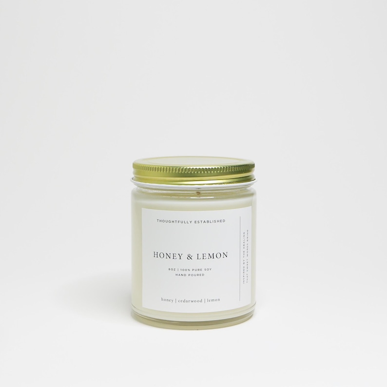 May include: A glass jar candle with a gold lid. The label reads "HONEY & LEMON" and "8oz | 100% PURE SOY | HAND POURED". The candle is white with a gold lid. The text "honey | cedarwood | lemon" is at the bottom of the label.