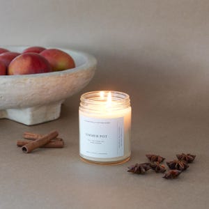 May include: A lit candle in a clear glass jar with a white label reading SIMMER POT, 100% PURE SOY, HAND POURED. Beside it are two cinnamon sticks and several star anise pods. In the background, a textured off-white bowl holds several red apples.
