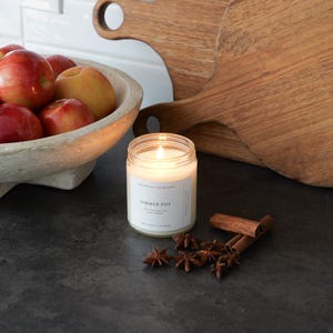 May include: A lit Simmer Pot candle in a clear glass jar with a white label. Cinnamon sticks and star anise are scattered around the candle. A wooden bowl of apples and two wooden cutting boards are in the background.