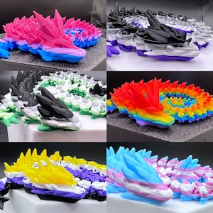 May include: A collection of 3D-printed dragon sculptures in various colors, each representing different LGBTQ+ pride flags. The dragons feature detailed, crystalline wings and bodies, with colors including pink, blue, purple, black, white, and rainbow hues.