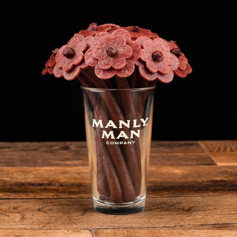 May include: A bouquet of cured meat flowers in a clear glass vase with the text "MANLY MAN COMPANY" printed on the side.