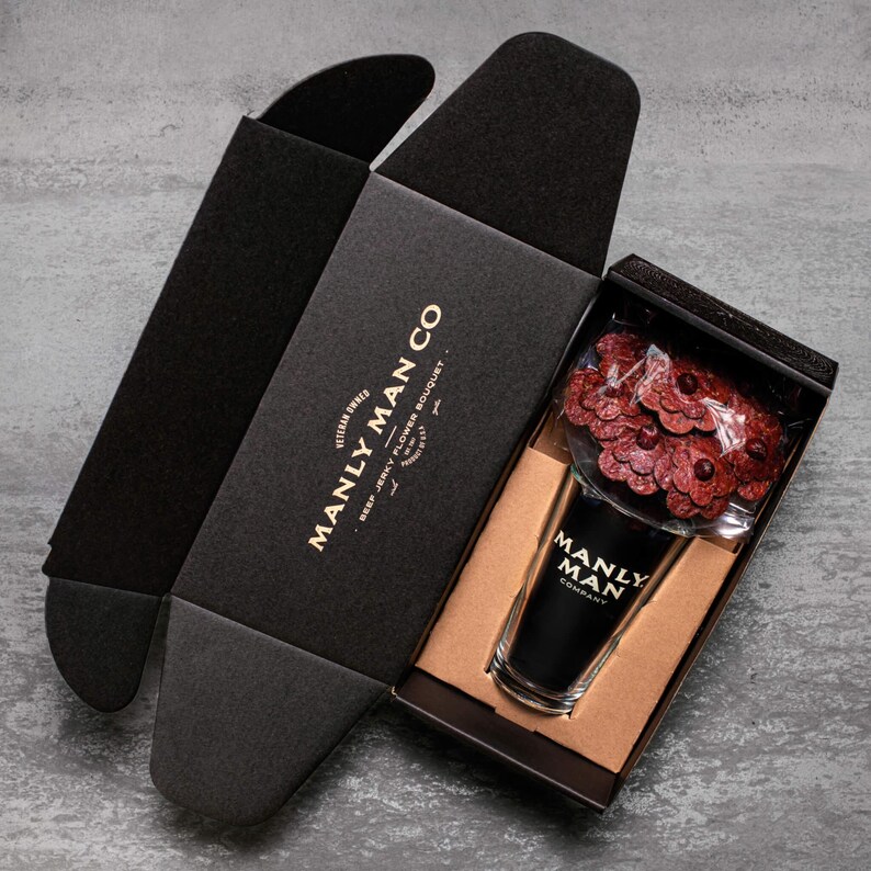 May include: A black box with the text "Manly Man Co.  Veteran Owned.  Beef Jerky Flower Bouquet" is open to reveal a black glass with a red and brown flower bouquet inside. The glass has the text "Manly Man Co." on it.