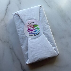 May include: A white book wrapped in white tissue paper with a round sticker that says "so many books so little time" with a stack of books and flowers in the center of the sticker.