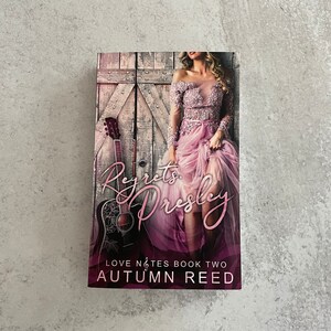 May include: Book cover with a woman in a pink dress standing in front of a wooden door with a guitar leaning against it. The title of the book is "Regret's Presley" and the author is "Autumn Reed". The book is part of a series called "Love Notes".