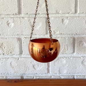 May include: A polished copper-coloured spherical hanging planter suspended by a thin gold-toned metal chain. The smooth, reflective surface of the bowl shows a distorted reflection of the surroundings, and the chain features small, uniform links.