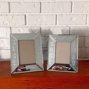 Peut inclure: Deux cadres photo rectangulaires en verre avec des motifs de feuilles gravées. Les cadres argentés sont posés sur une surface en bois devant un mur de briques blanches. Les cadres sont vides.