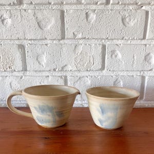 May include: Two handmade ceramic pieces: a small jug with a handle and a small bowl. Both are beige with blue watercolour-like accents. The pieces sit on a wooden surface against a white brick wall.