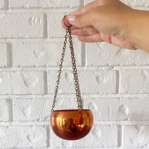 May include: A small, spherical, metallic copper-colored hanging planter with a smooth, reflective surface. The bowl is suspended by three thin, matching copper-toned metal chains that connect to a single circular ring at the top for hanging.