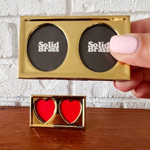 May include: A rectangular gold-toned brass double picture frame featuring two oval openings with black inserts labeled Solid Brass. Below it sits a smaller rectangular gold-toned brass double frame featuring two heart-shaped openings with red inserts.