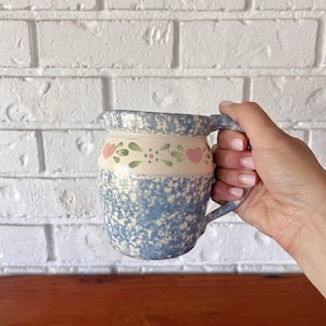 May include: A blue and white speckled ceramic jug with a handle. The jug features a cream-coloured band with pink hearts and green leaves. The jug is held in a hand.