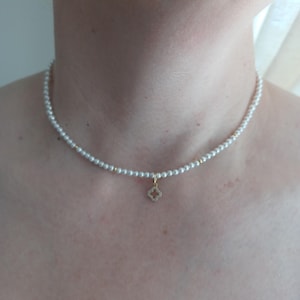 May include: A delicate choker necklace featuring a single strand of small white pearls interspersed with tiny gold-toned beads. A small, four-petaled clover-shaped pendant with a gold-toned border and embedded clear stones hangs from the center.