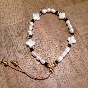 May include: A white and black beaded bracelet with gold accents and four white clover charms. The bracelet has a brown cord tie closure.