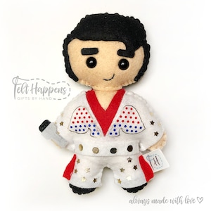 May include: Felt doll of Elvis Presley wearing a white jumpsuit with gold stars, red and blue wings, and a red scarf. He is holding a microphone and has a black pompadour hairstyle.