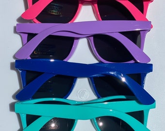 May include: A stack of sunglasses in various colors: red, pink, purple, blue, teal, and black. Each pair has dark lenses and a classic frame design. The sunglasses are made of polycarbonate and offer 100% UV protection.
