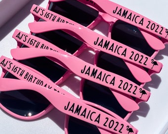 May include: Pink plastic sunglasses with black lenses. The sunglasses have the text "JAMAICA 2022" and a flamingo printed on the arms.