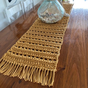 Op de afbeelding: Een gehaakte tafelloper met een franje rand in een mosterdgele kleur. De loper ligt op een houten tafel met een glazen vaas gevuld met schelpen.