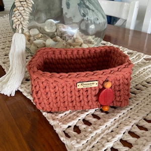 Könnte beinhalten: Ein rechteckiger, terrakottafarbener Häkelkorb mit einem strukturierten Stichmuster. Ein kleines Holzschild mit dem Text "Rosemade QUEENSLAND" ist angebracht. Eine dekorative Holzperle und -scheibe hängen an der Seite. Der Korb steht auf einem cremefarbenen, offen gewebten Tischset.