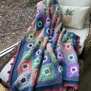 May include: A handmade crochet blanket in a patchwork design, featuring squares of various pastel colors including pink, blue, green, and purple. The blanket is draped over a wicker chair with a white cushion and a pineapple-themed pillow.