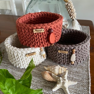 May include: Three handmade crochet baskets in terracotta, cream, and grey. Each basket has a wooden tag that reads "Rosemade Queensland" and decorative wooden beads. The baskets are displayed on a grey patterned surface with seashells and greenery.