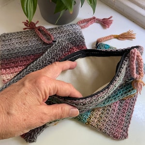 May include: Two crocheted zippered pouches with a pink, grey, and teal colour scheme. The pouches are open and the lining is visible.
