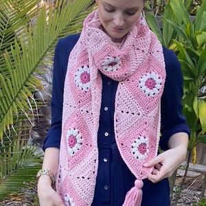 May include: A pink and white crochet scarf with a floral pattern and tassel ends. The scarf is made of soft yarn and features a repeating diamond design with white and pink flower motifs. The scarf is approximately 72 inches long.