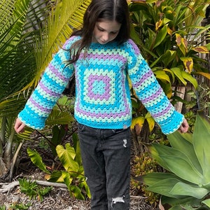 May include: A crocheted sweater with a square pattern in shades of blue, green, and purple. The sweater has long sleeves and a relaxed fit.