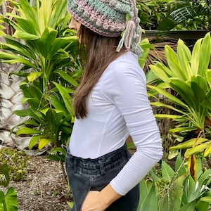 May include: A crocheted hat with a long tassel in shades of pink, green, and white. The hat is worn by a person with long brown hair and a white long-sleeved shirt.
