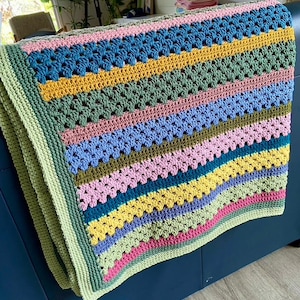 May include: A colorful, handmade crochet blanket with a striped pattern. The blanket features rows of various colors, including pink, blue, yellow, green, and teal. The edges are finished with a light green border. The blanket is folded over a dark blue surface.