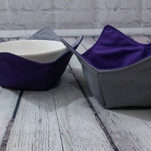 May include: Two fabric bowl cozies, one purple and one gray, with a white ceramic bowl inside one of the cozies.