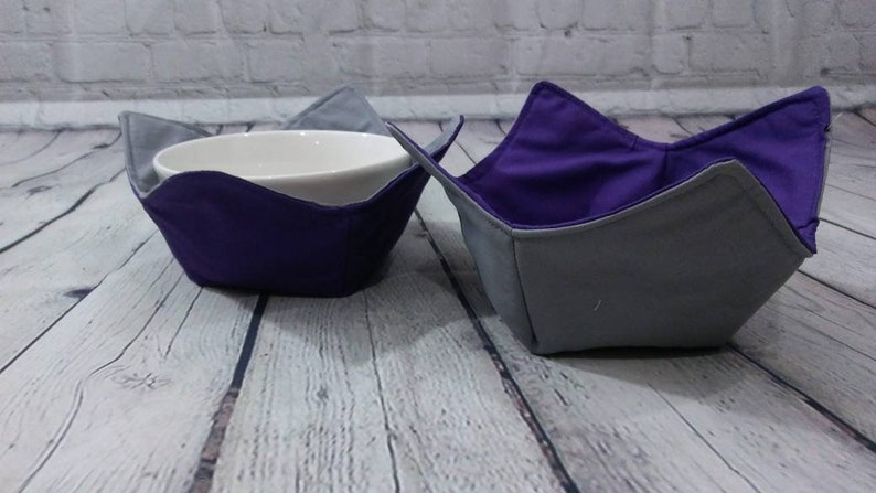 May include: Two fabric bowl cozies, one purple and one gray, with a white ceramic bowl inside one of the cozies.