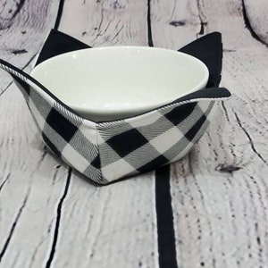 May include: A white ceramic bowl sits inside a black and white buffalo check bowl cozy with black cat ears. The cozy is designed to hold the bowl and keep the contents warm. The background is a white wooden surface.