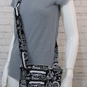 May include: Black crossbody bag with a repeating white text design. The bag features a long strap and multiple zippered compartments. The text includes words like "Jesus", "Peace", and "Lamb of God". The bag is displayed on a mannequin.