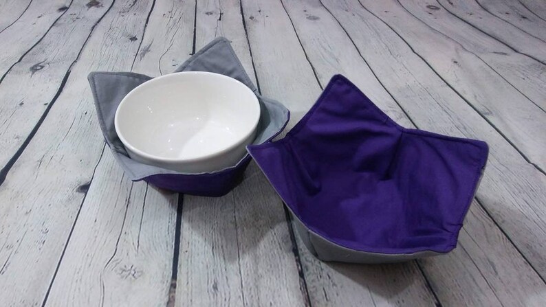 May include: Two fabric bowl cozies, one gray and one purple, with a white ceramic bowl inside the gray cozy.