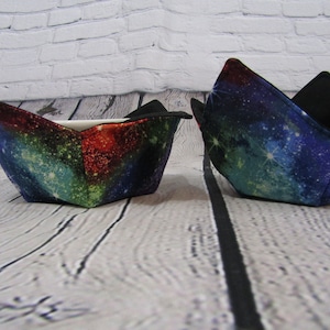 May include: Two fabric bowl cozies with a galaxy print in shades of blue, green, red, and black. The cozies are folded and sitting on a white wooden surface.