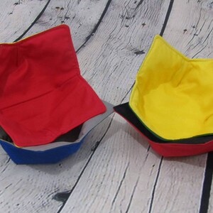 May include: Three fabric bowl cozies in red, yellow, and blue. The cozies are made of a soft, quilted fabric and are designed to protect bowls from heat and scratches.