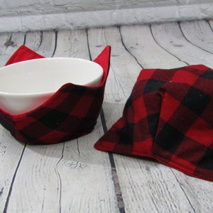 May include: Two red and black plaid bowl cozies. One cozy is holding a white bowl. The cozies are made of a soft, quilted fabric.