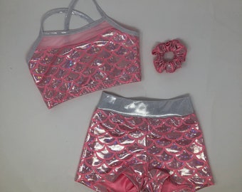 May include: A pink mermaid-scale patterned dance set, including a crop top, shorts, and scrunchie. The top has a silver trim and mesh detail. The shorts have a silver waistband. The scrunchie matches the set. The set is designed for dance or gymnastics.