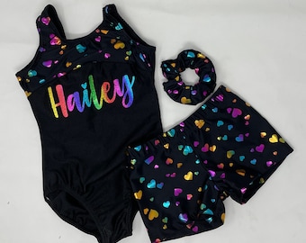 Peut inclure: Ensemble noir comprenant un justaucorps, un short et un chouchou, ornés de cœurs multicolores. Le justau-corps porte le nom "Hailey" en lettres arc-en-ciel. Idéal pour la gymnastique ou la danse.
